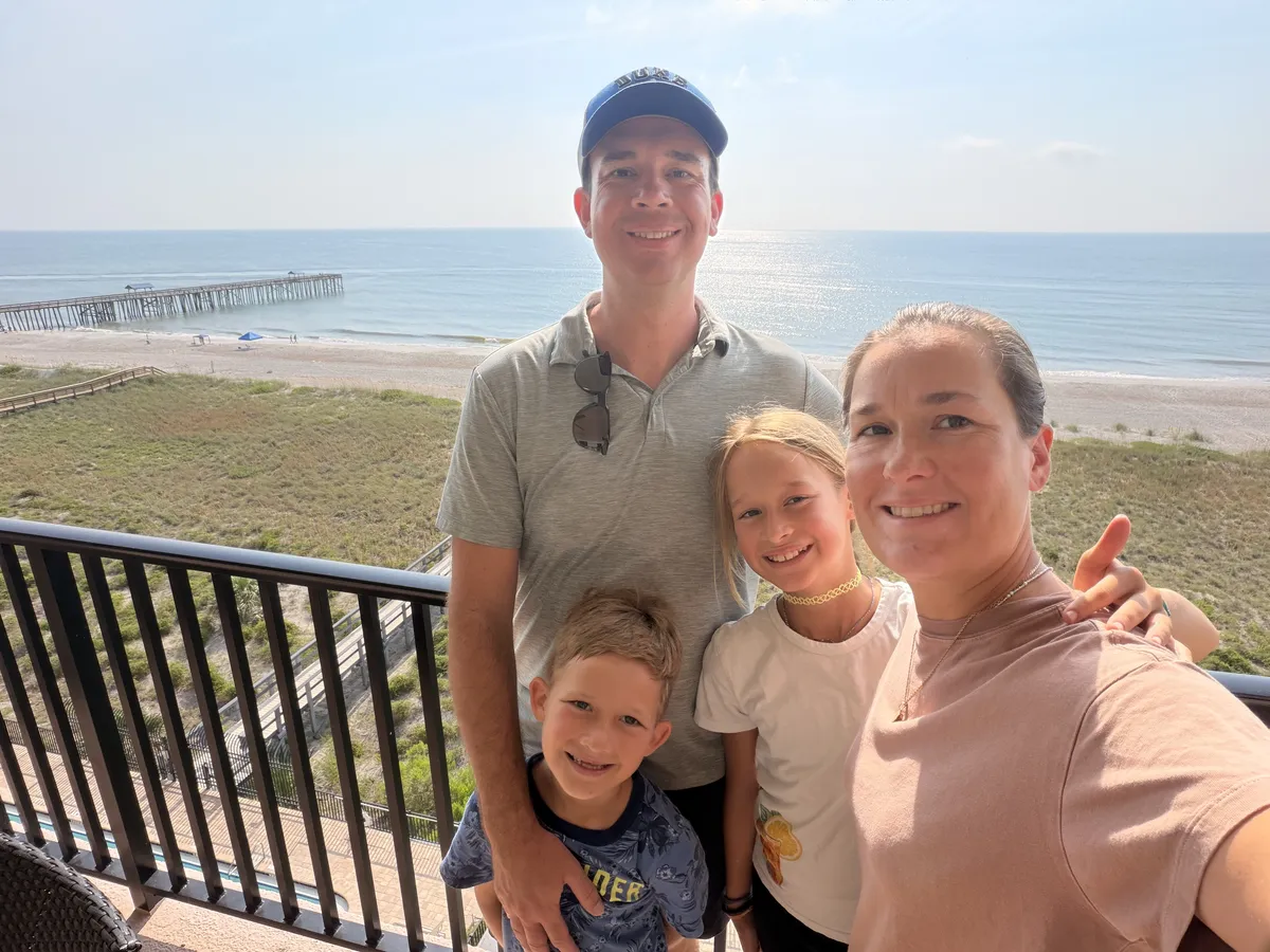 A family of four smiling on a balcony above the beach